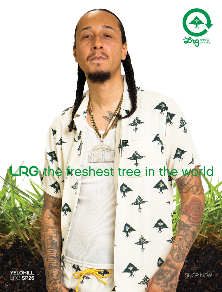 Man with braided hair wearing a patterned shirt with LRG branding, surrounded by grass.