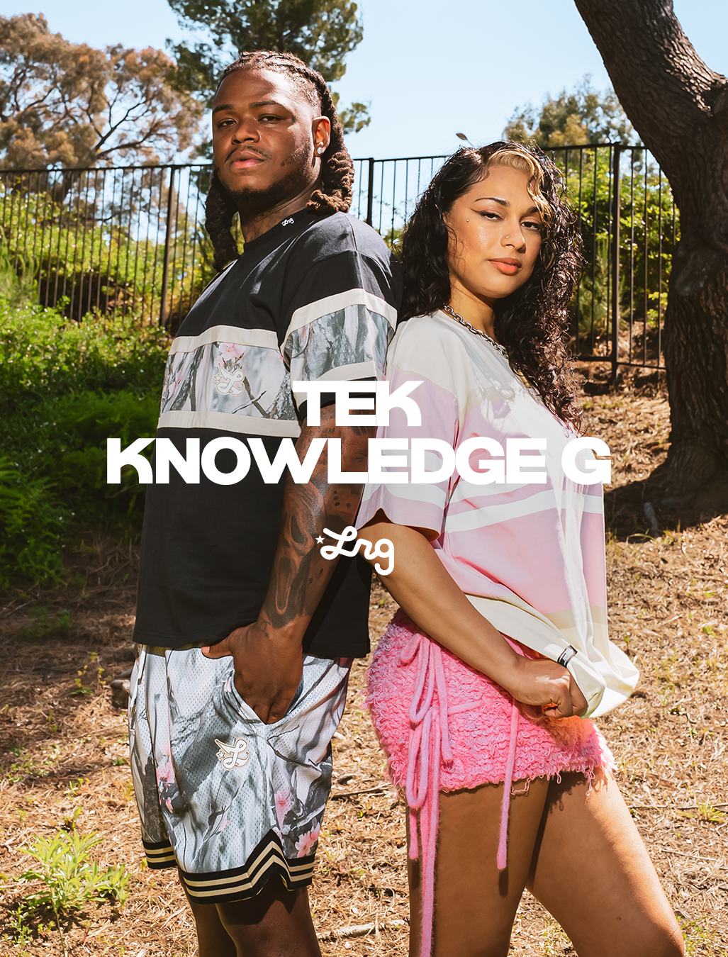 Two models standing outdoors in a sunny park-like setting, wearing oversized striped graphic t-shirts and shorts. The man wears a black and gray striped shirt with gray patterned shorts, and the woman wears a white and pink striped shirt with a pink fringed skirt.